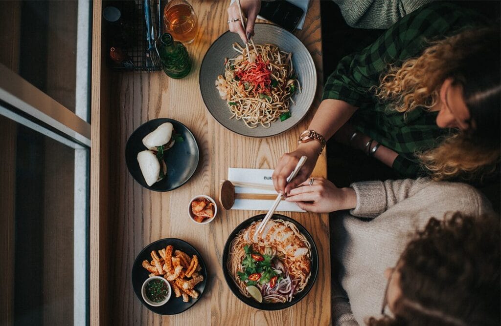 Two people sit at a wooden table by a window, enjoying Asian-inspired dishes from The Restaurant Group, including noodles, bao buns, kimchi, and seasoned fries. Drinks and condiments are also on the table as they use chopsticks to eat. MHP Group discuss supporting the move from AIM to FTSE 250.