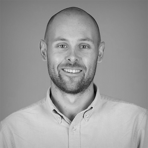 A man with a shaved head and short beard, wearing a light-colored button-up shirt, smiles at the camera against a plain, neutral background. The image is in black and white.