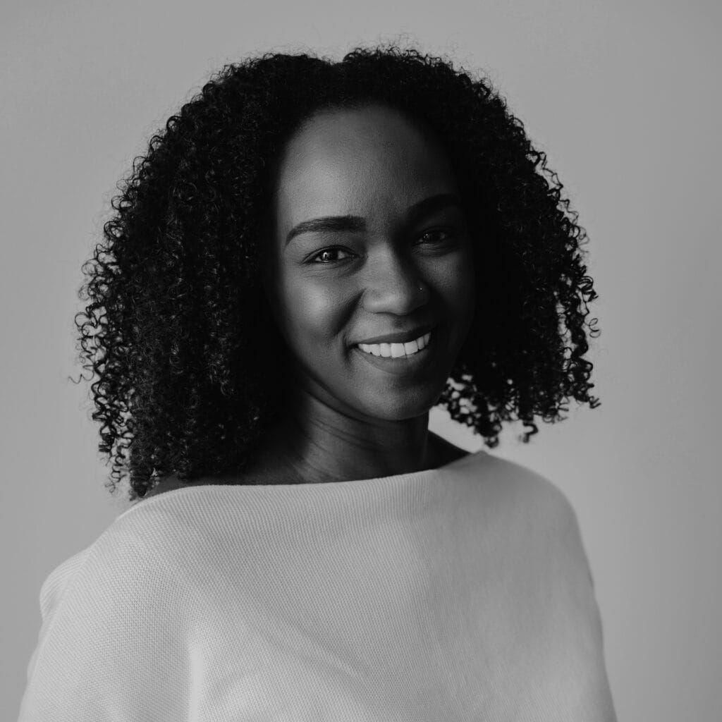 Raneisha, a woman with curly hair, smiles warmly at the camera. She is wearing a light-colored top and is photographed in black and white against a plain background.