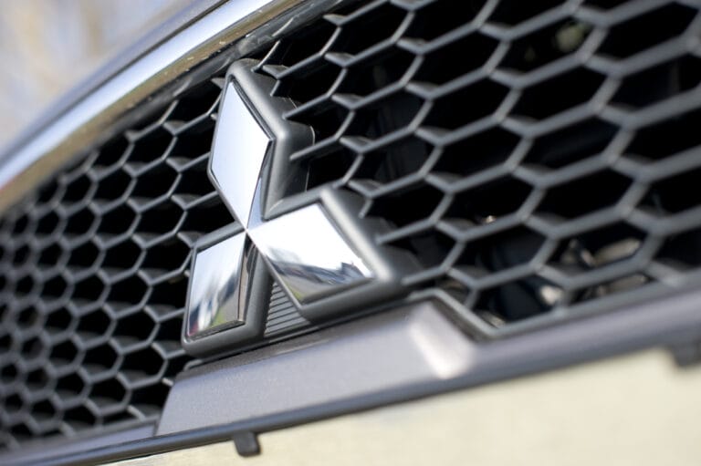 Close-up of a Mitsubishi car grille featuring the chrome Mitsubishi Electric logo with a honeycomb pattern background, highlighting sleek design and innovative EMEA Strategic Communications.