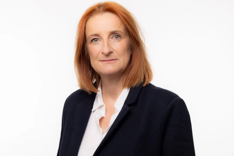 Mirror Editor-In-Chief Alison Phillips, a middle-aged woman with straight, shoulder-length red hair, wears a black blazer over a white shirt and stands against a plain white background.