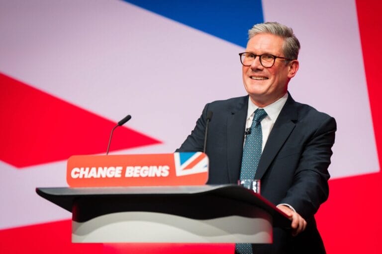 A political insider in a suit and glasses smiles while standing at a podium labeled CHANGE BEGINS in front of a large red, white, and blue background.