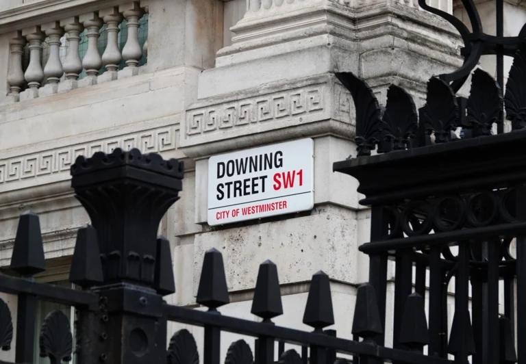 A street sign on a stone building reads “Downing Street SW1, City of Westminster,” partially obscured by an ornate black metal fence—a striking scene in a city where low trust voters look to Labour and anti-establishment coalitions for change.