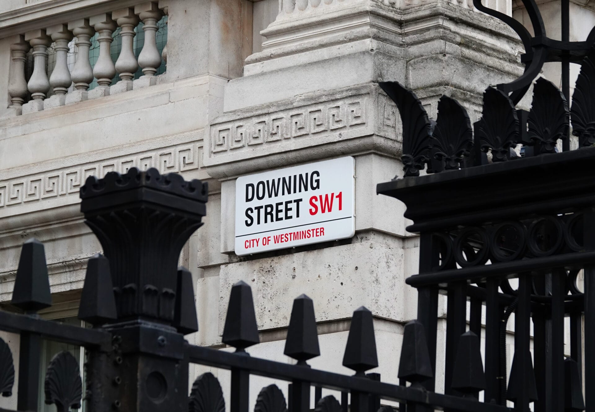 A street sign on a stone building reads “Downing Street SW1, City of Westminster,” partially obscured by an ornate black metal fence—a striking scene in a city where low trust voters look to Labour and anti-establishment coalitions for change.