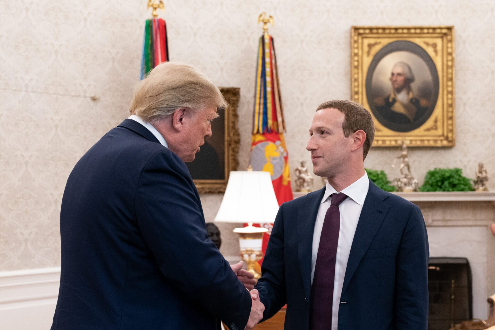 Two men in suits shake hands in an ornate room with flags, a lamp, and a framed portrait of George Washington, highlighting the trust built during high-stakes discussions.