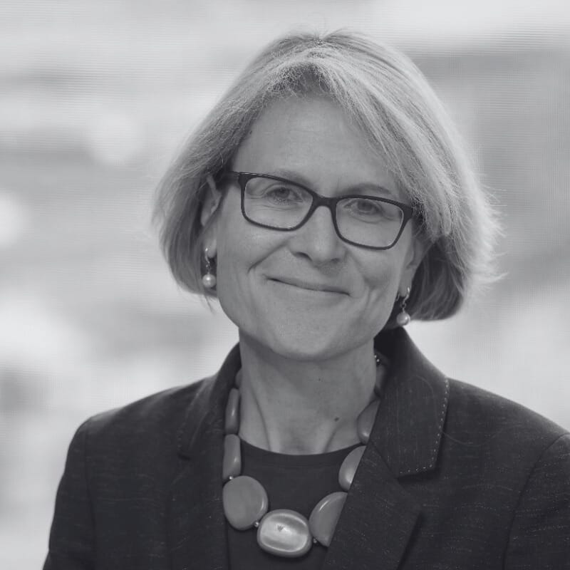 Victoria Wakely, a woman with short, light hair and glasses, smiles at the camera. She is wearing a dark blazer, a beaded necklace, and earrings. The background is softly blurred.
