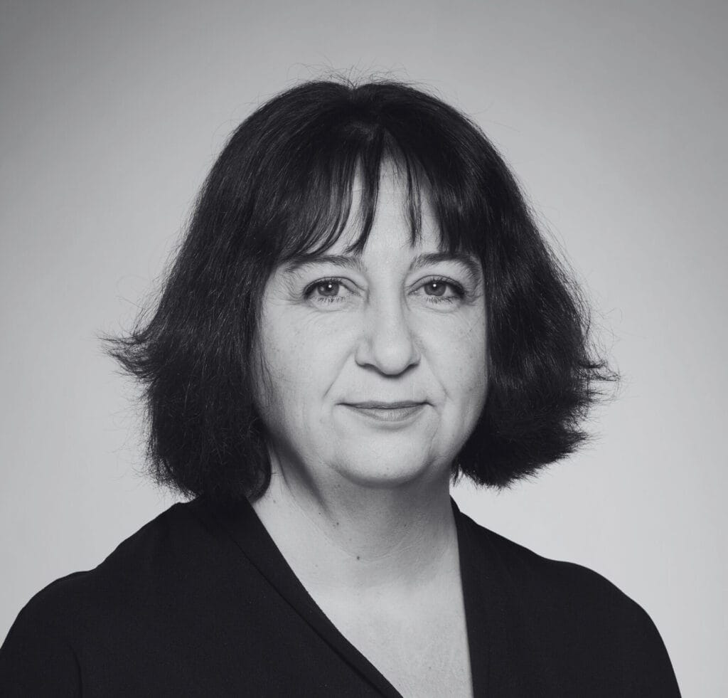 Black and white portrait of Clare Wilson, a middle-aged woman with medium-length dark hair and bangs, wearing a dark top, facing the camera with a neutral expression against a plain background.