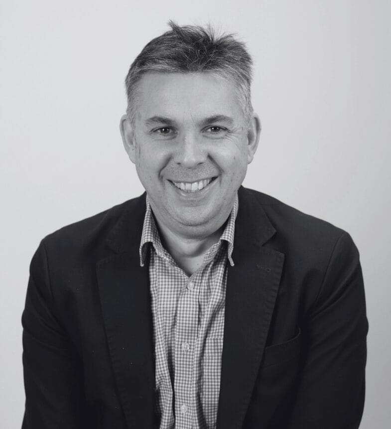 A man with short hair smiles at the camera, wearing a checked shirt and a dark blazer. The background is plain and light-colored. The image is in black and white.