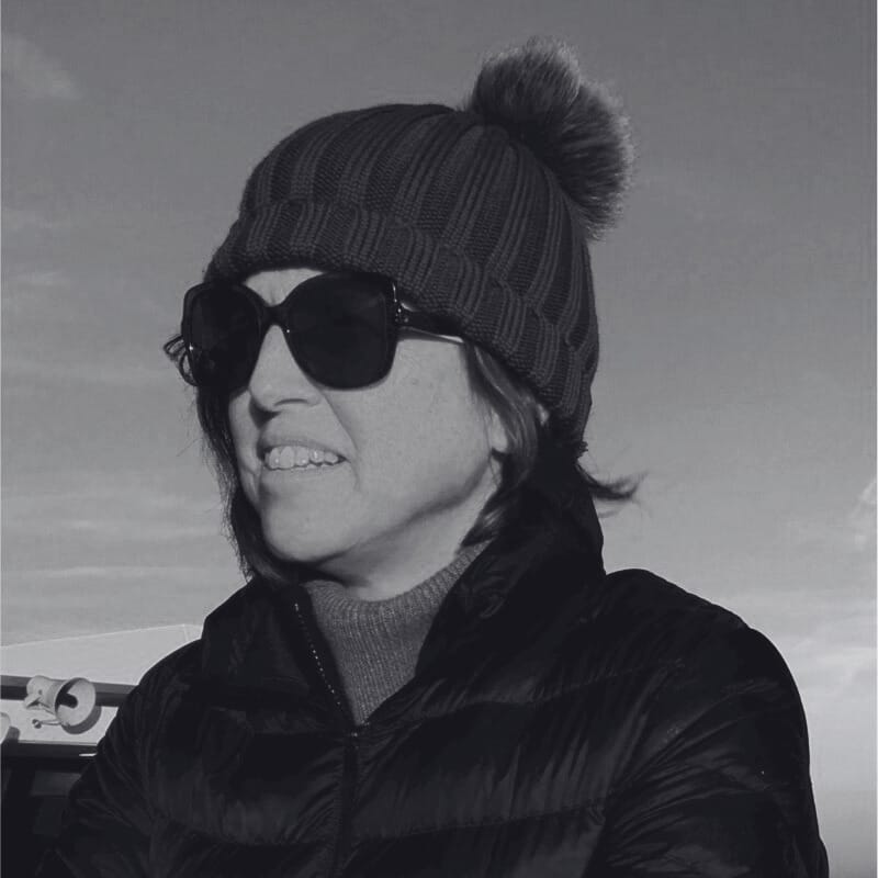 Sarah Whitehead, wearing a knit beanie with a pom-pom, sunglasses, and a puffy jacket, smiles outdoors on a cold day. The image is in black and white.
