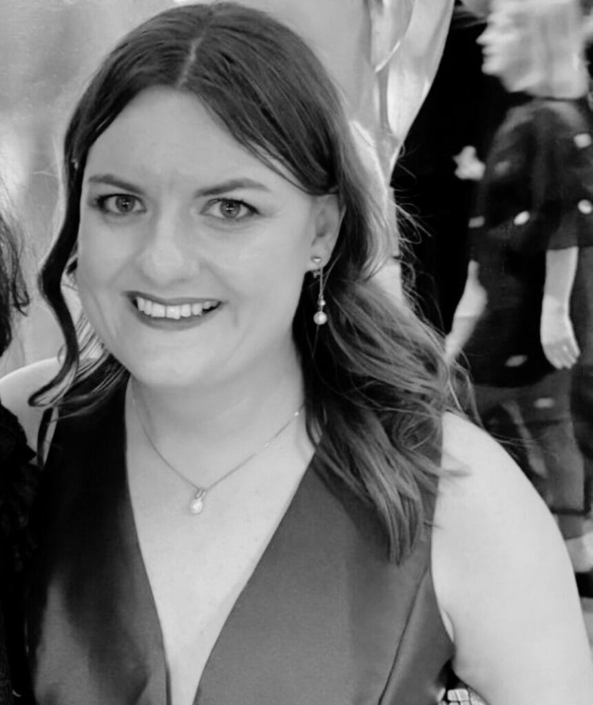 Geraldine McKelvie, with wavy hair, wearing a sleeveless V-neck dress and drop earrings, smiles at the camera. Other people are visible in the blurred background. The image is in black and white.