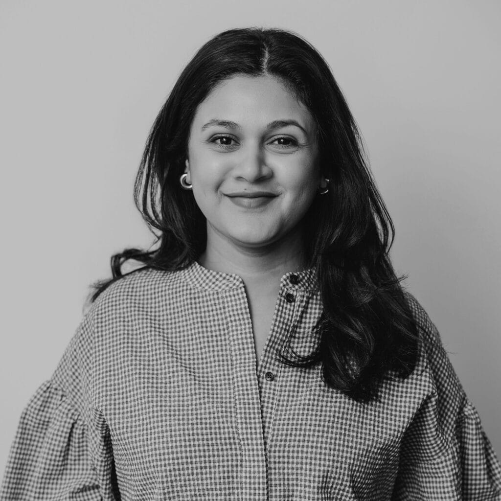 Aditi Karnad, a woman with long dark hair, wearing a checked blouse with buttons, smiles softly while facing the camera against a plain background. The image is in black and white.