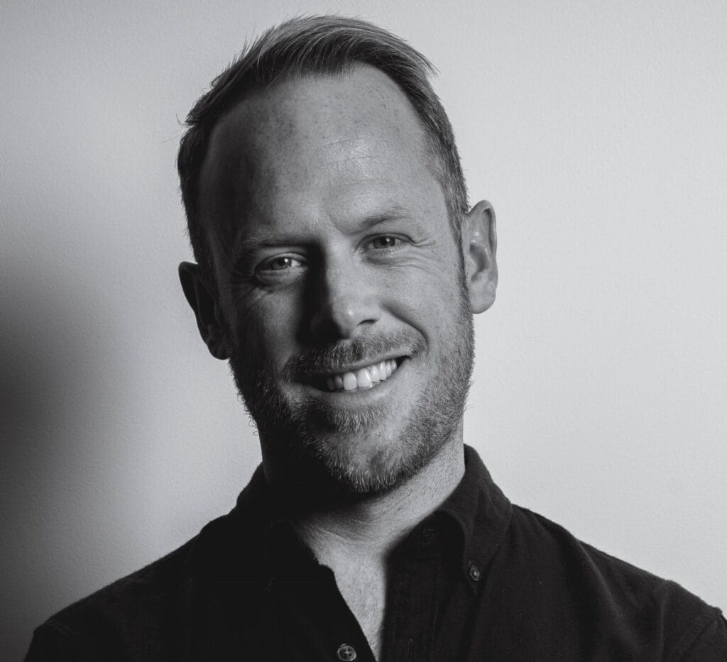 Black and white portrait of James Shapland, a smiling man with short hair and a beard, wearing a button-up shirt, standing against a plain light-colored background.