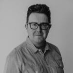 Black and white portrait of a man with short, dark hair and glasses, wearing a button-up shirt. He is facing the camera and smiling slightly, with a plain light background.