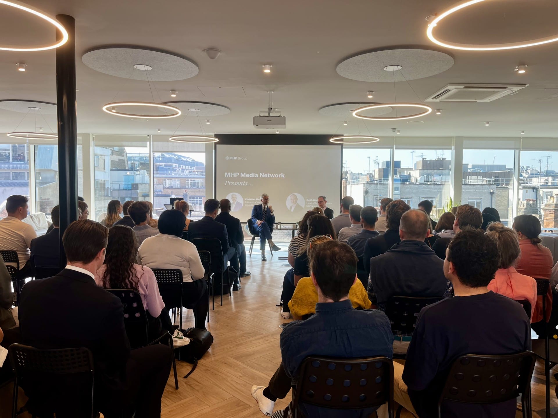 A group of people seated in a modern, bright conference room listens to three speakers at the front, including City AM Editor in Chief Christian May, with a presentation projected on a screen and large windows revealing an urban cityscape.