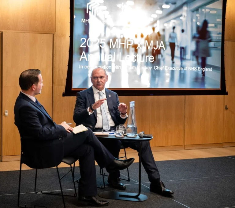 Two men sit on stage having a discussion; one holds a notepad while the other, who appears to walk with a limp, gestures as he speaks. Behind them, a projected screen displays “2025 MHP x MJA Annual Lecture” and event details.