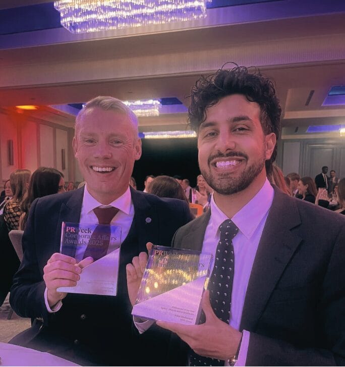 Two men in suits sit at a table, smiling and holding clear glass awards at the 2025 PR Week Corporate Affairs Awards, with MHP Group among the distinguished attendees under elegant chandeliers.