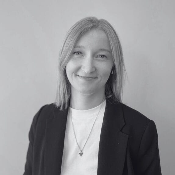 A person with straight, shoulder-length hair is smiling at the camera. They are wearing a dark blazer over a light shirt and a necklace with a small pendant. The background is plain and light-colored. The image is in black and white.