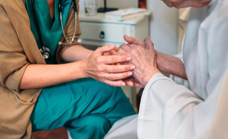 A healthcare worker in scrubs gently holds the hands of an elderly person, offering comfort and support in a clinical setting focused on compassionate end-of-life care.