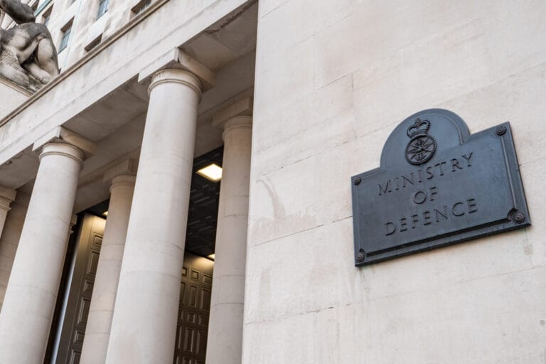Close-up of the Ministry of Defence building’s stone facade, featuring four tall columns, an open door, and a black plaque with the Ministry of Defence emblem and text, highlighting its pivotal role in shaping UK defence strategy.