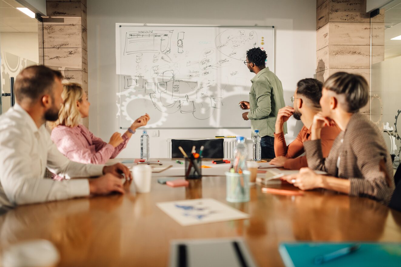 A man stands at a whiteboard covered with diagrams and notes, giving a presentation on brand strategy to four colleagues seated around a conference table in a modern office.
