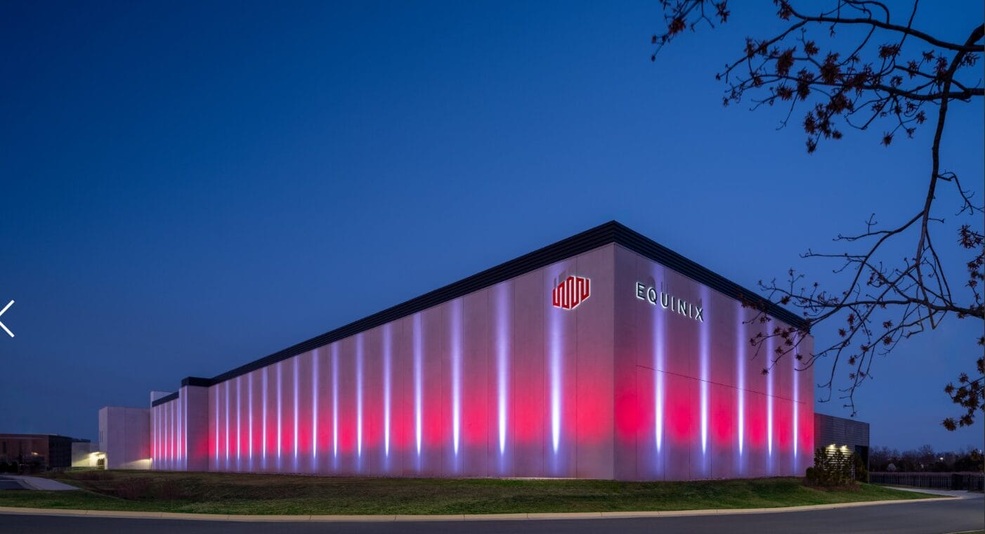 A large, modern Equinix data center building is lit with red and violet lights at dusk. The Equinix logo and name are visible on the illuminated exterior wall, providing a striking corporate brief. A tree and empty road are in the foreground.