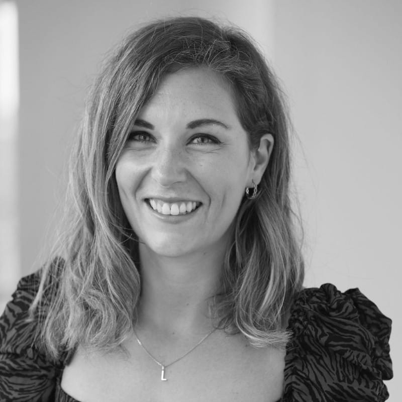 Black and white photo of Lesley Reddihough, a woman with wavy shoulder-length hair, smiling. She is wearing a patterned top, small hoop earrings, and a necklace with an L pendant. The background is plain and softly lit.
