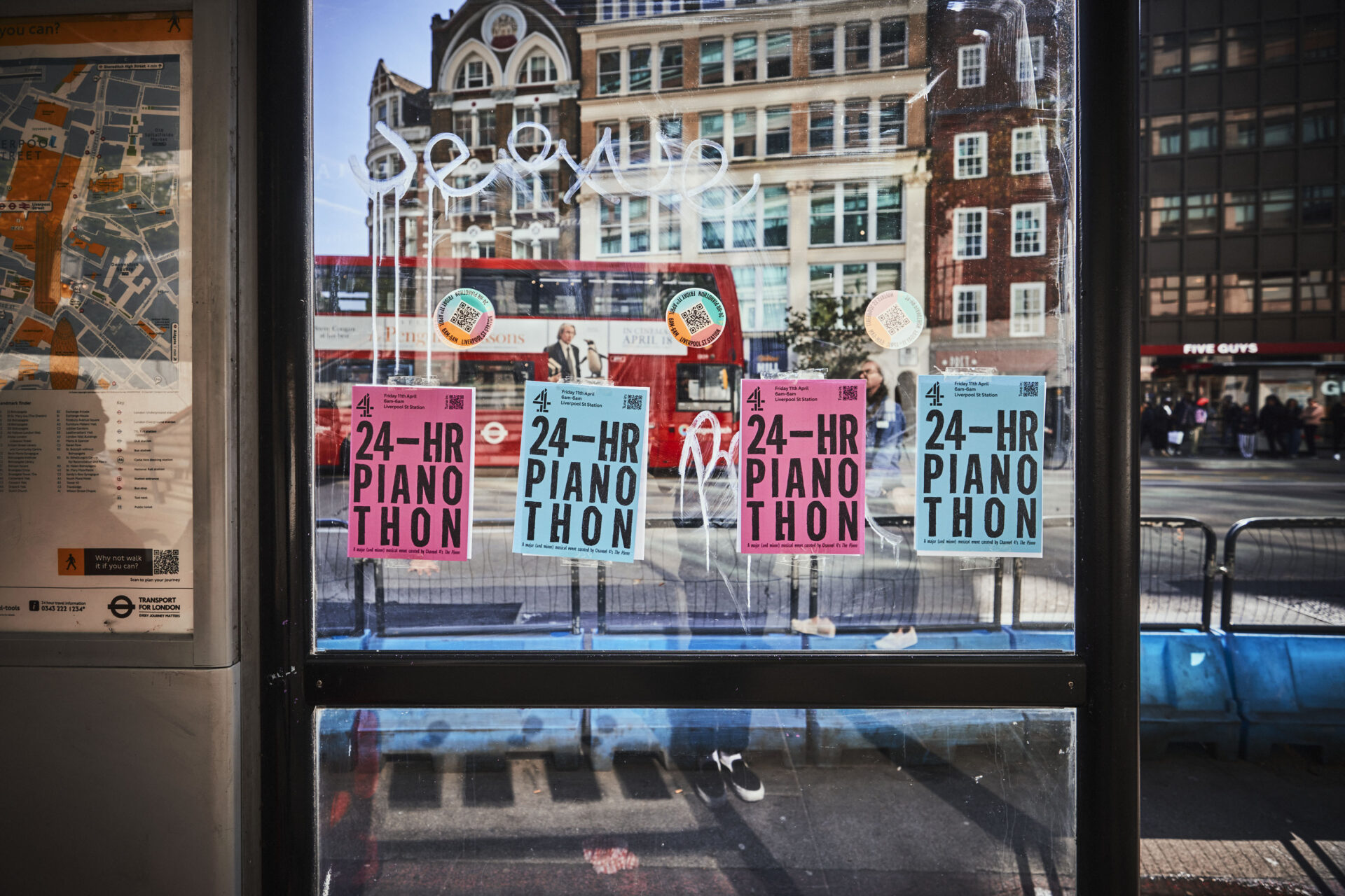 Posters for a 24-hour Piano Thon are taped to the glass of a bus stop shelter, with a city street, buildings, and a red double-decker bus visible in the background.