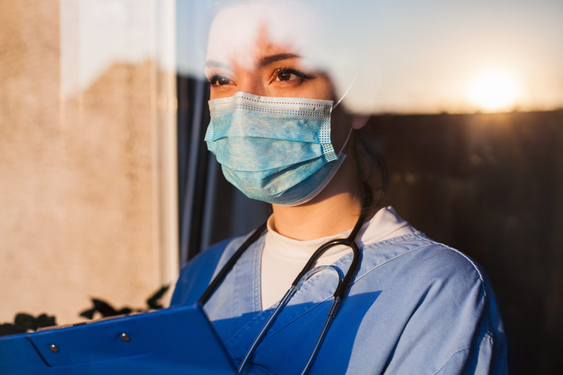 A healthcare worker wearing a surgical mask and scrubs stands by a window, holding a clipboard—a true test of dedication as sunlight shines through the glass, partially reflecting her face.