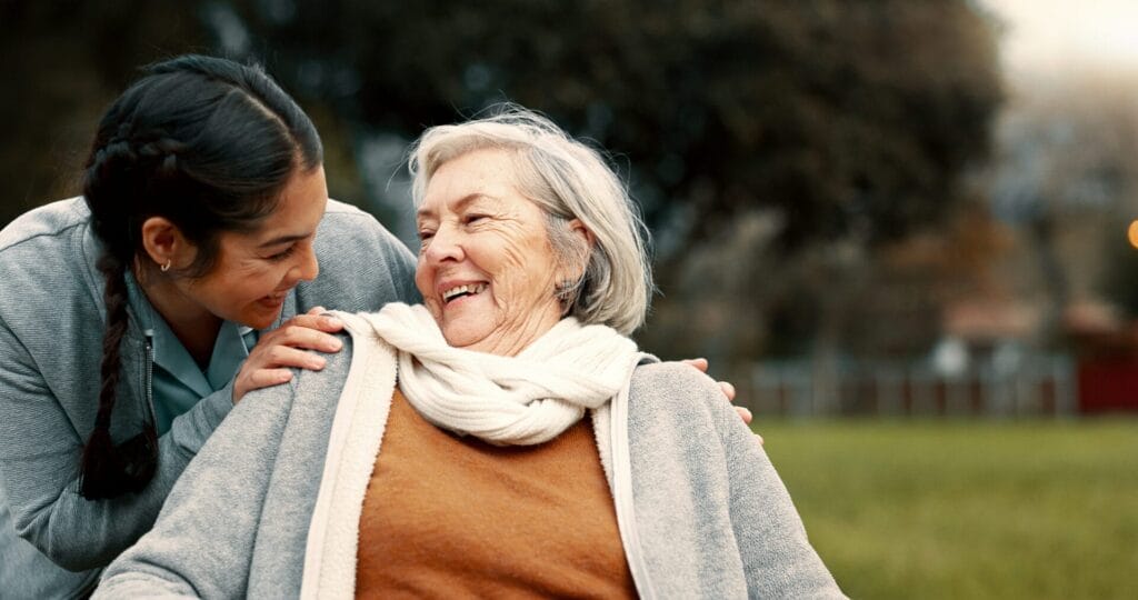 A smiling elderly woman in a wheelchair wearing a scarf is outdoors, laughing with a younger woman who is leaning close and touching her shoulder, both radiating the joy and warmth that defines our Masterbrand differentiation.