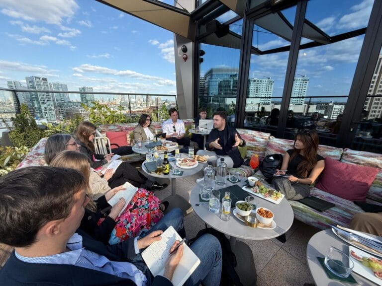 A group of people sit around tables on a rooftop terrace, engaged in discussion about AI. There are drinks, notebooks, and snacks on the tables. Tall city buildings and a blue sky with clouds are visible in the background.