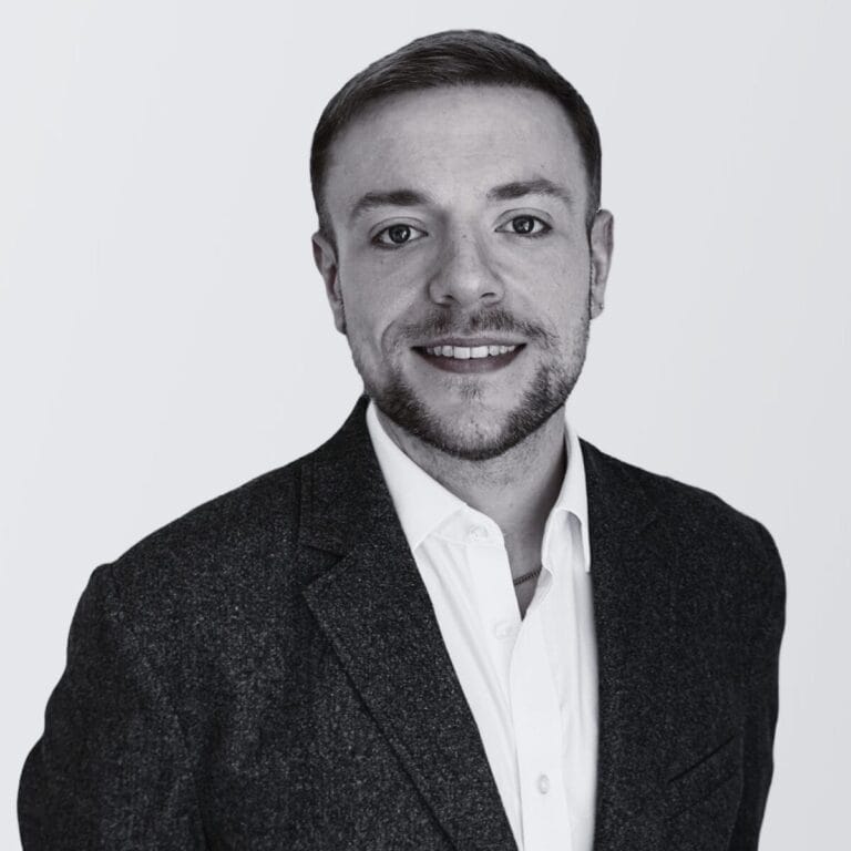 Stefan Roberts, a man with short hair and a trimmed beard, wearing a dark blazer over a white shirt, smiles at the camera against a plain light background.